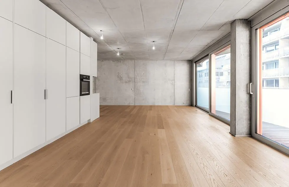 An empty room with light wood floors, white cabinets, and a concrete wall and ceiling. Sliding glass doors lead to a balcony.