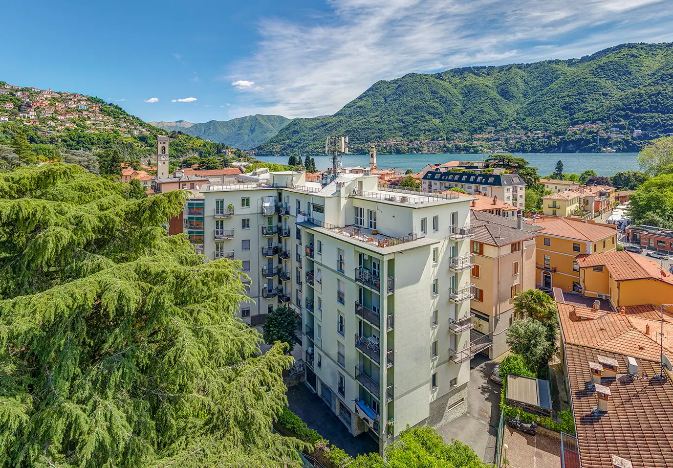 Scenic view of a light-colored apartment building with balconies, overlooking Lake Como, Italy, with mountains in the background.