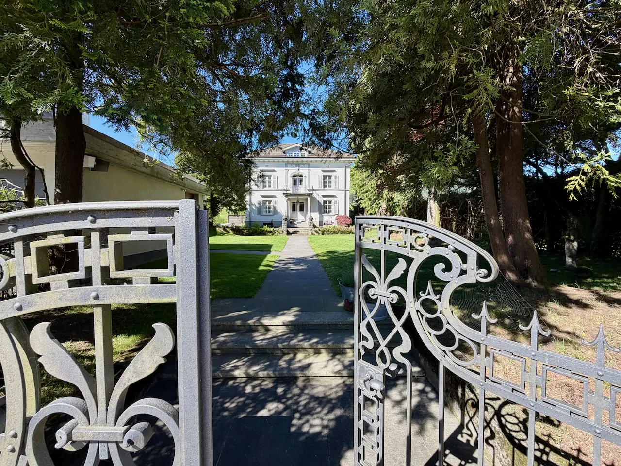 View through ornate metal gates to a white, two-story house with green lawn and trees.