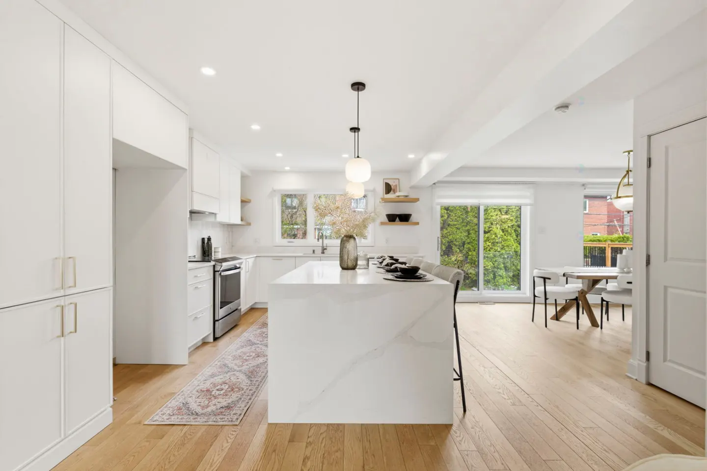 Bright, modern kitchen with white cabinets, marble island, wood floors, and dining area visible through sliding glass doors.