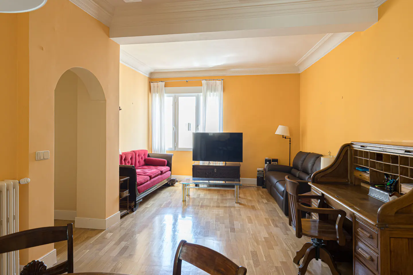 Living room with orange walls, hardwood floors, a red sofa, a black leather sofa, a TV, and a wooden roll-top desk.