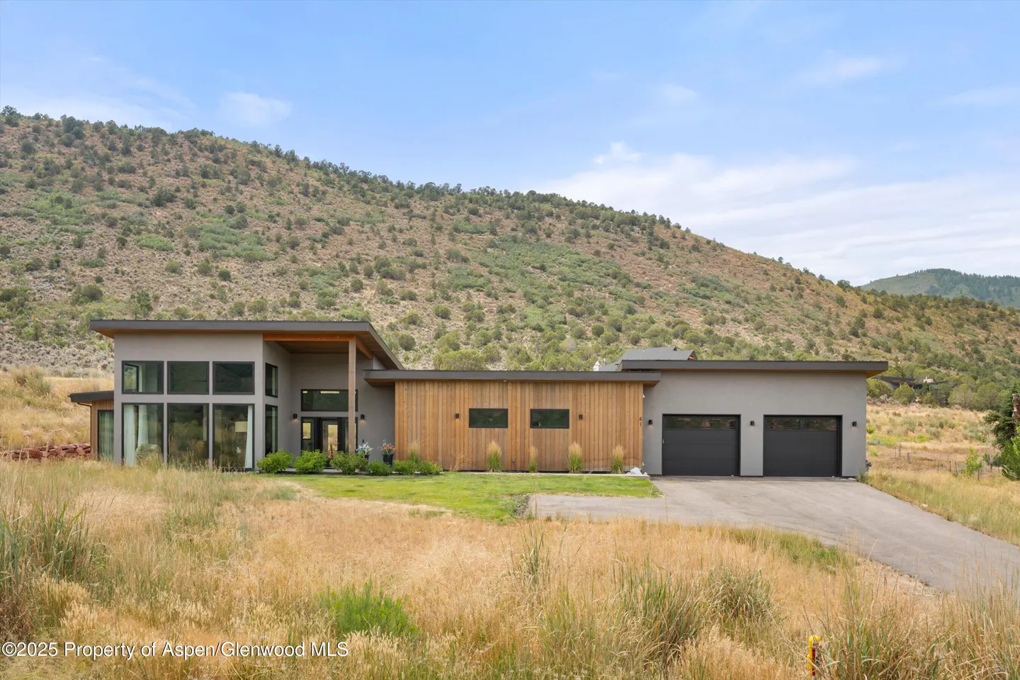 Modern home with gray and wood exterior, large windows, and a two-car garage. A mountain rises in the background under a blue sky.