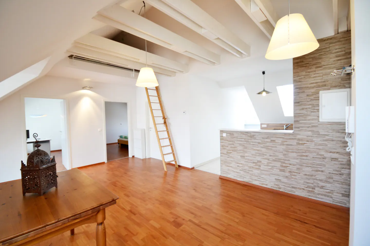Bright, open-plan attic apartment with wooden floors, white walls, and exposed beams. A ladder leans against the wall, and pendant lights hang from the ceiling.