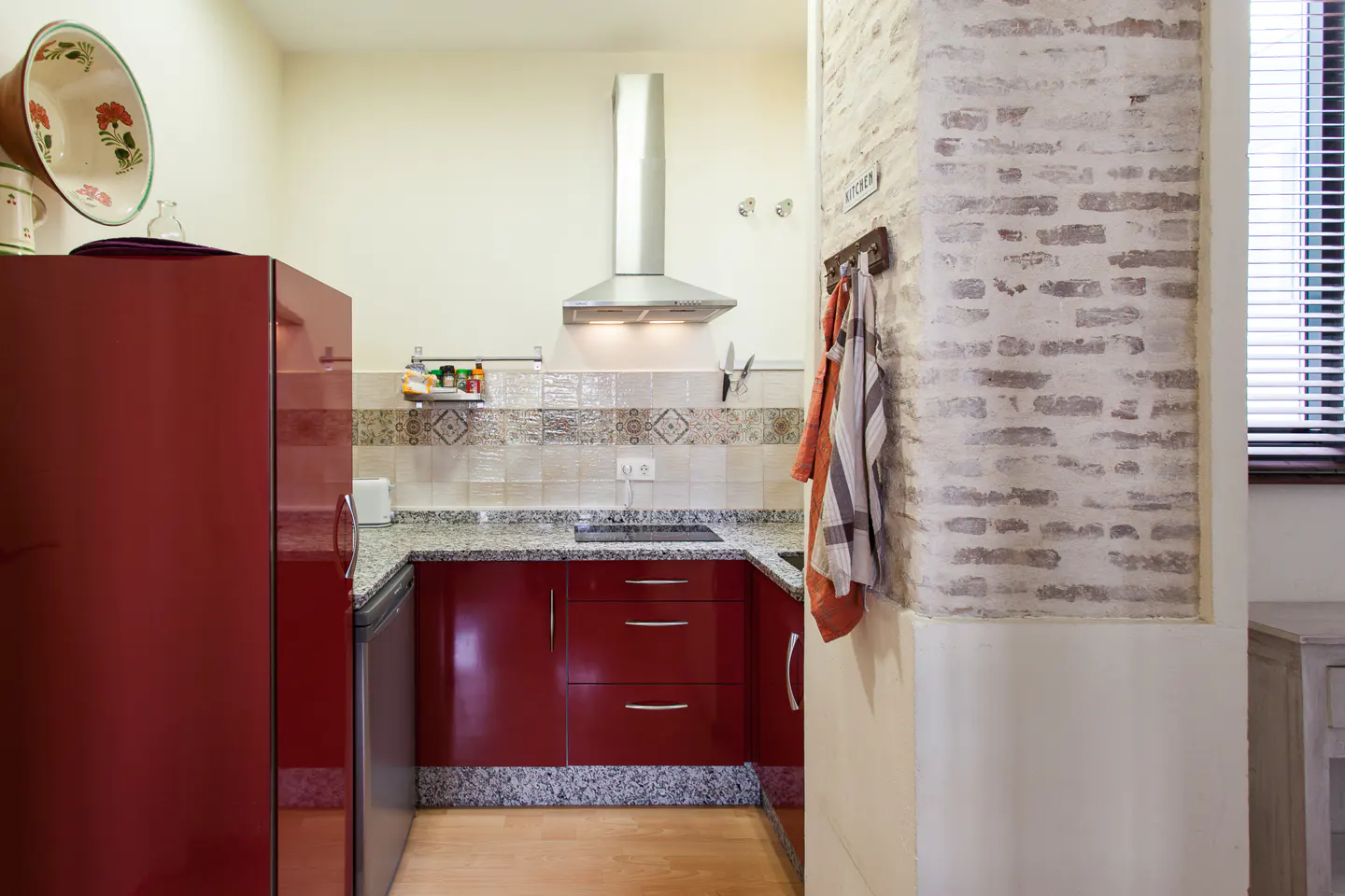 A compact kitchen with red cabinets, a stainless steel range hood, and a red refrigerator. A brick-style wall is on the right.