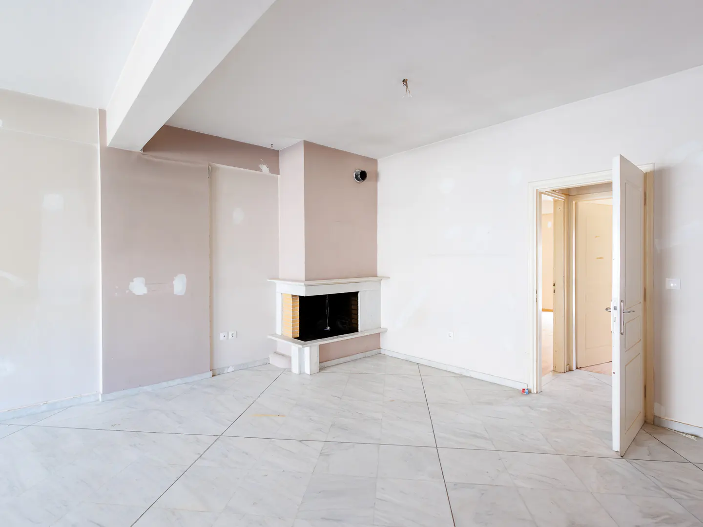 Empty room with white marble floors, a corner fireplace, and white walls under renovation, leading to an open doorway.