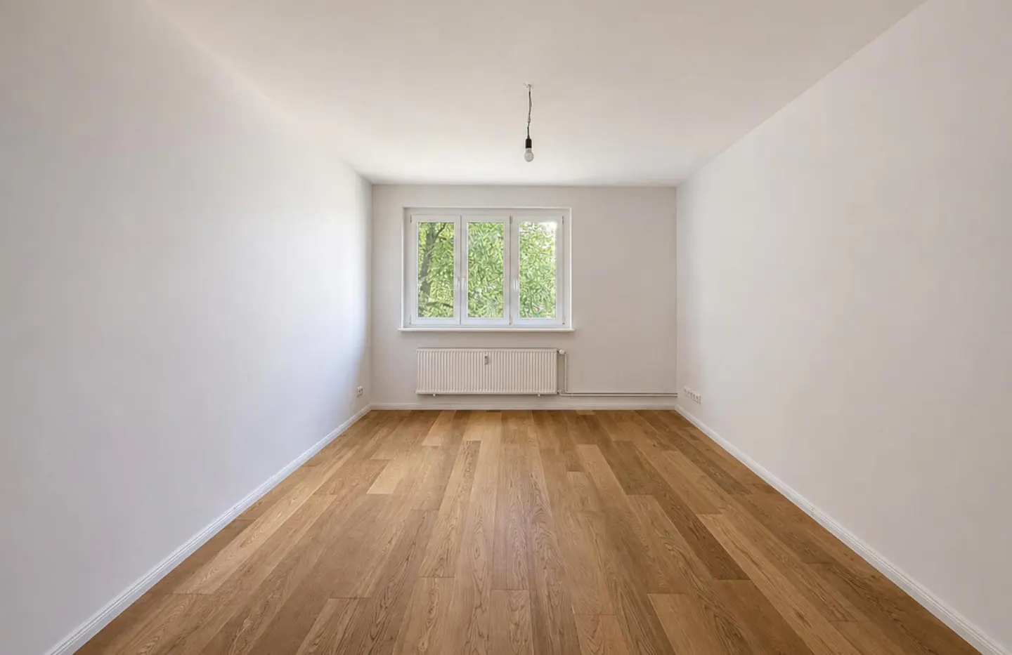 Empty room with white walls, light wood flooring, a central window with a view of trees, and a white radiator below.