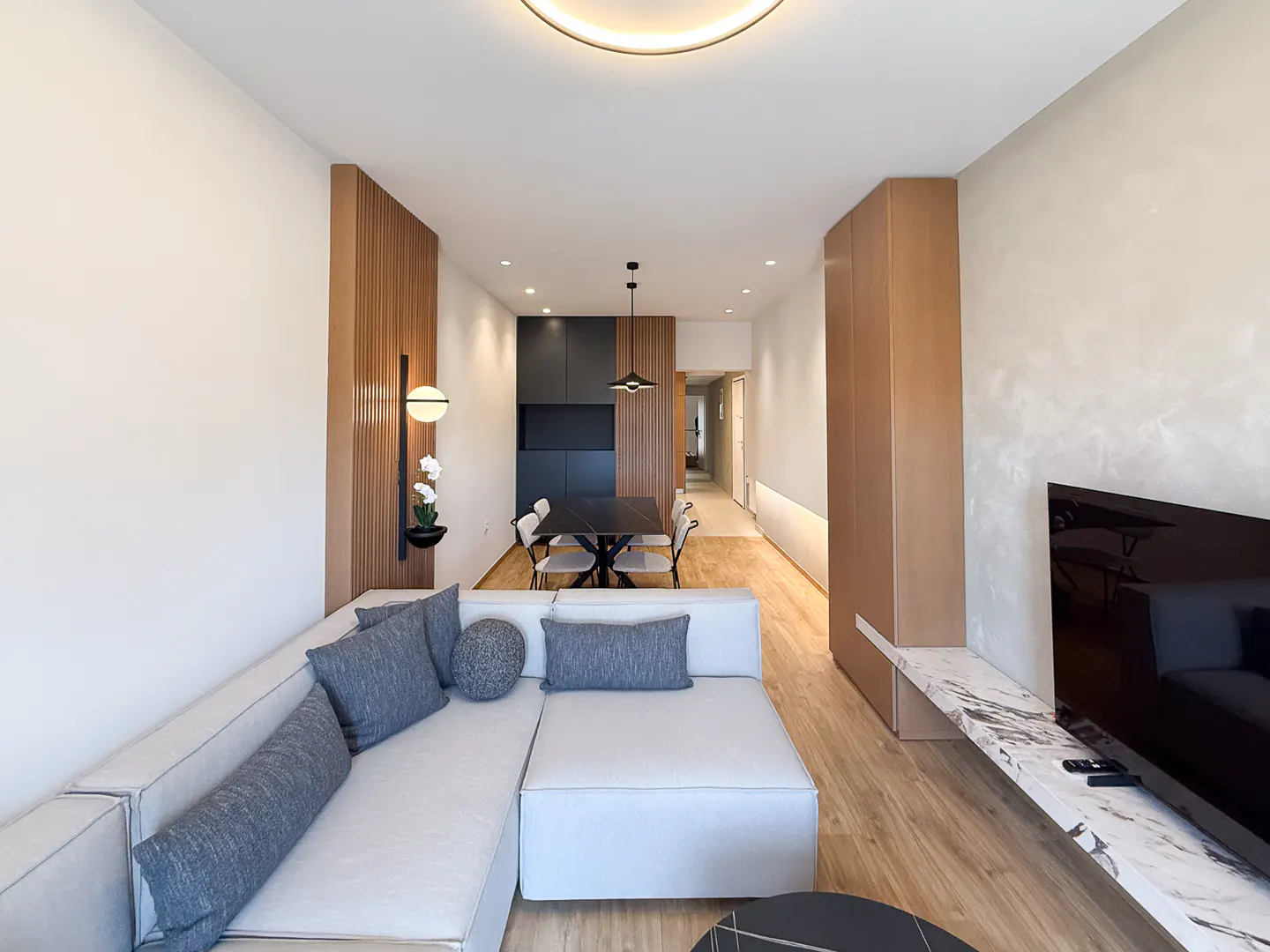 Bright, modern living room with a white sectional sofa, wood floors, and a black dining table with white chairs. A large TV is mounted on the wall.