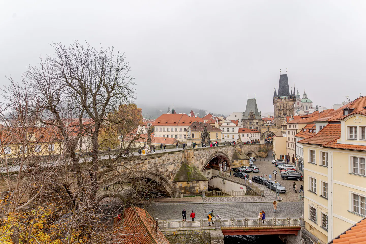 A high-angle view of Prague’s Charles Bridge with stone arches, historic buildings, and people walking on a cloudy day.