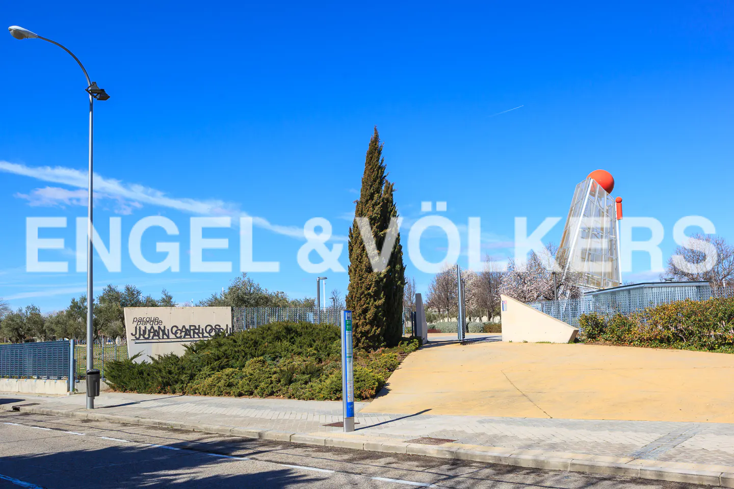 Park entrance with blue sky. A tall tree stands near a bus stop. In the background is a sculpture with an orange sphere.
