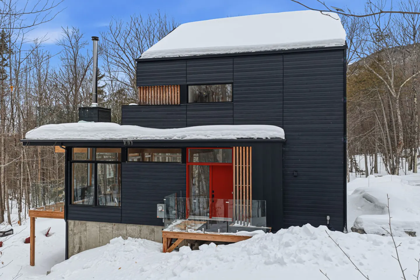 Modern black house with a red door, surrounded by snow and trees. The roof is covered in snow.