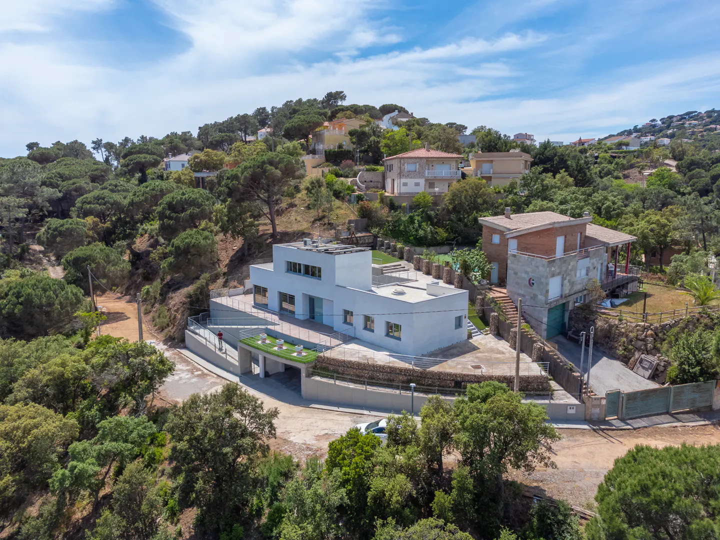 Aerial view of a modern, white, flat-roofed house nestled on a green hillside with other homes and trees under a blue sky.