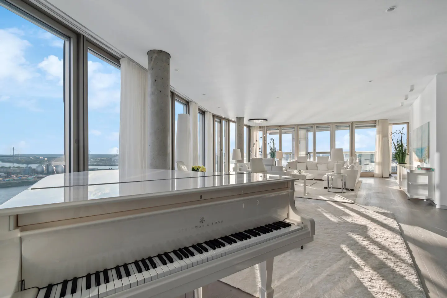 Bright, modern living room with a white grand piano, white furniture, and floor-to-ceiling windows overlooking a cityscape and river.