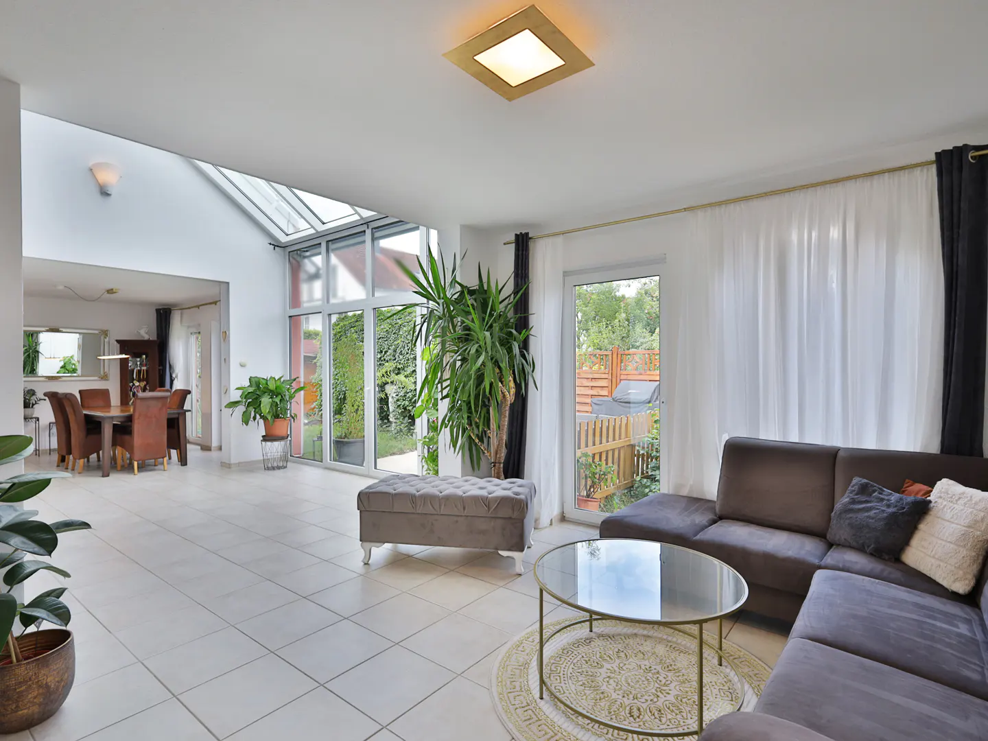 Bright, open-concept living room with tile floors, brown sectional sofa, glass coffee table, and dining area in the background.