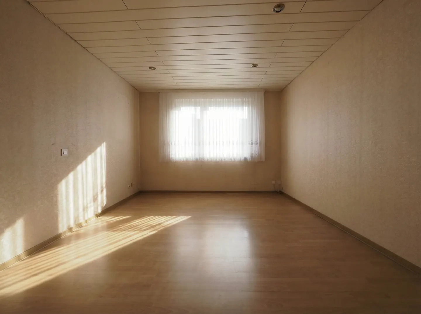 Empty room with light wood floors, beige walls, and a white wood plank ceiling. A window with sheer white curtains is centered on the back wall. Sunlight streams in.