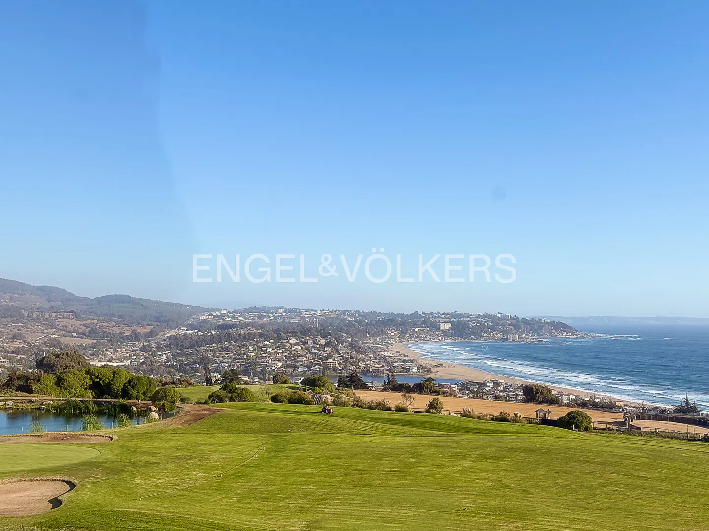 Scenic view of a green golf course overlooking a coastal town and the ocean under a clear blue sky.