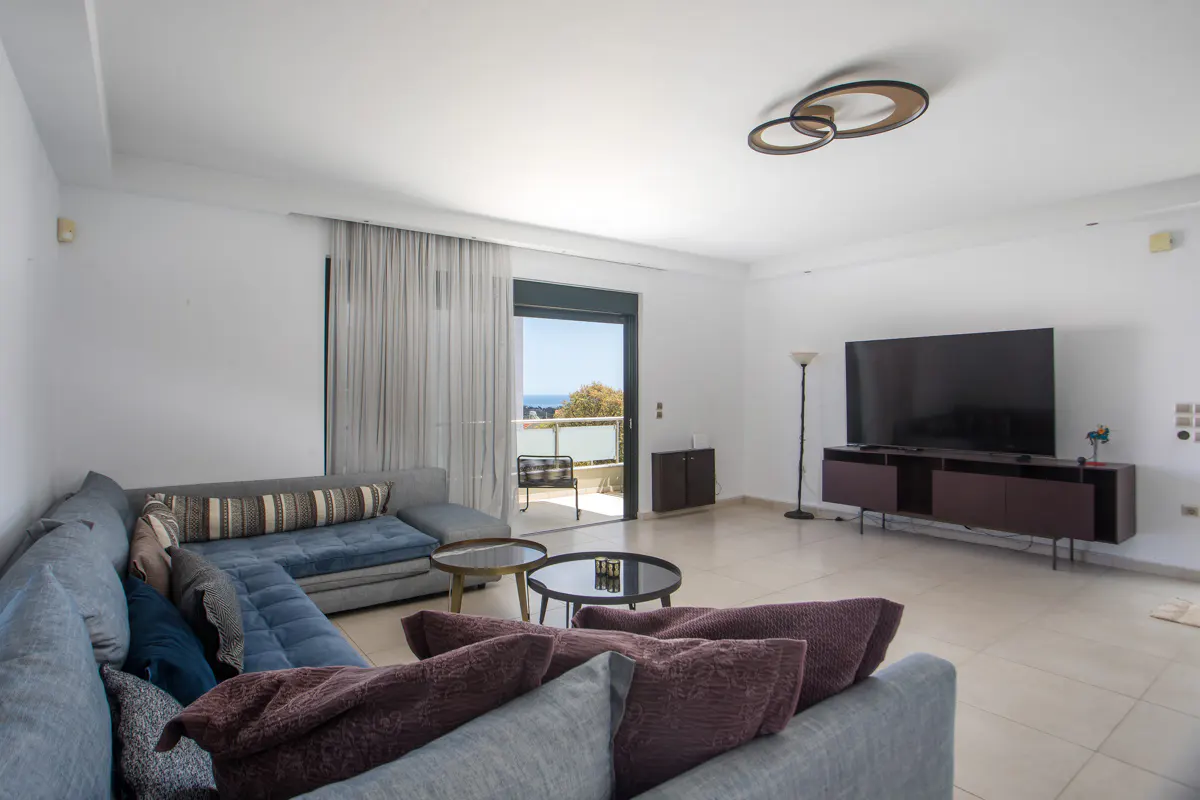 Bright living room with gray sofas, brown pillows, and a TV. A balcony with ocean views is visible through sliding glass doors.