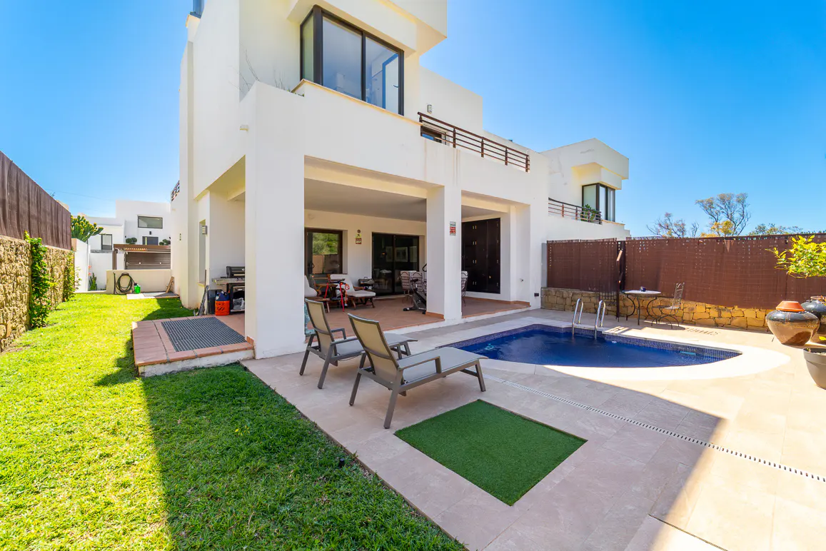 Backyard of a modern white house with a blue pool, lounge chairs, green grass, and a covered patio on a sunny day.