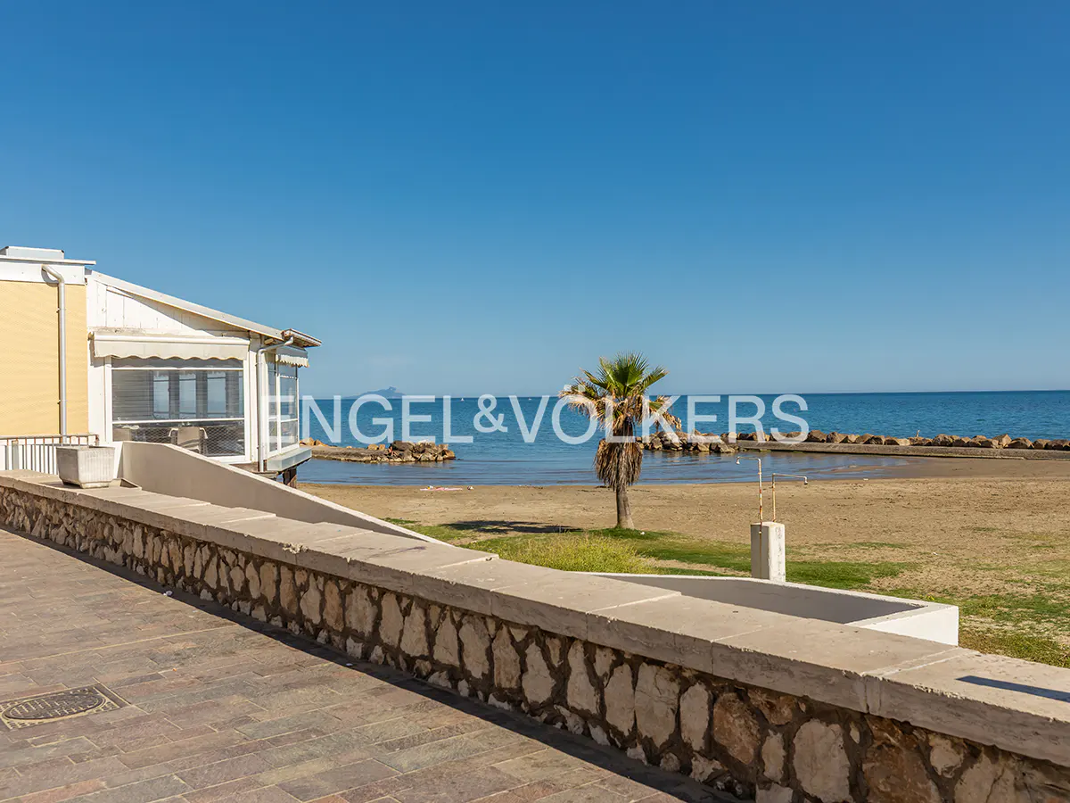 Beachfront property with a yellow building, stone wall, palm tree, and ocean view under a clear blue sky.