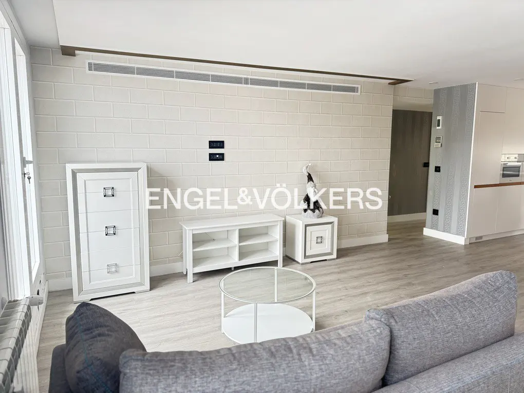 Modern living room with light brick wall, white furniture, glass coffee table, and a grey sofa in the foreground.