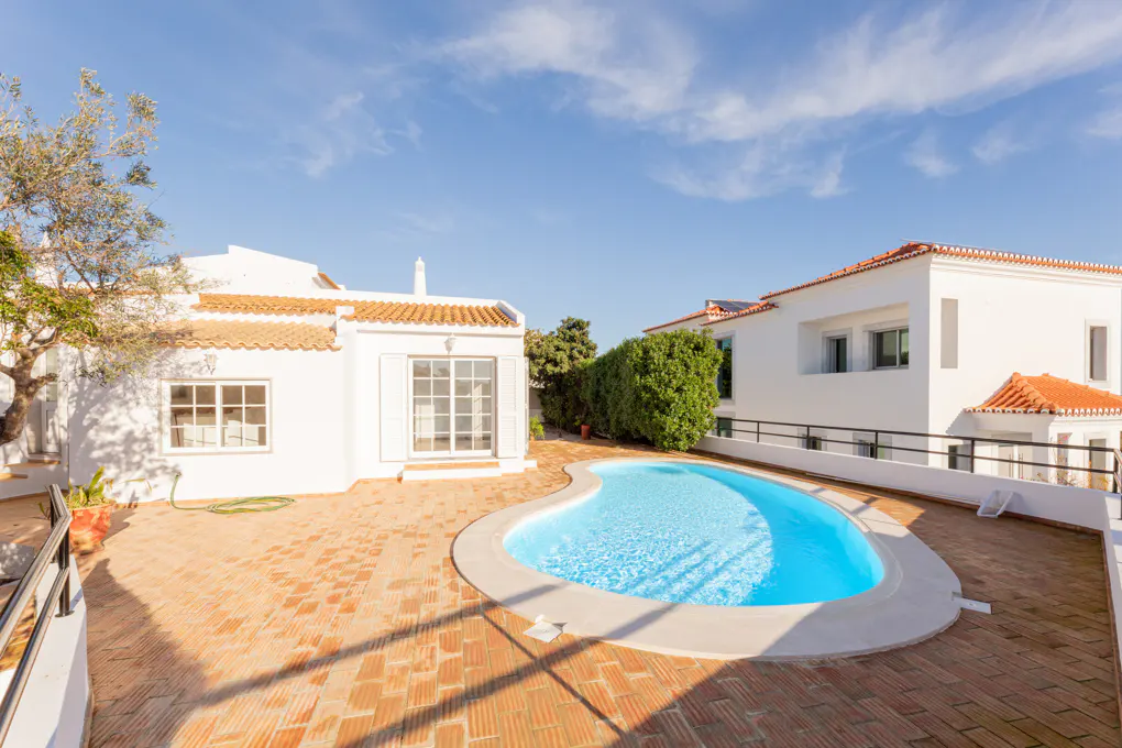 Exterior view of a white house with a red tile roof, a blue kidney-shaped pool, and a brick patio on a sunny day.