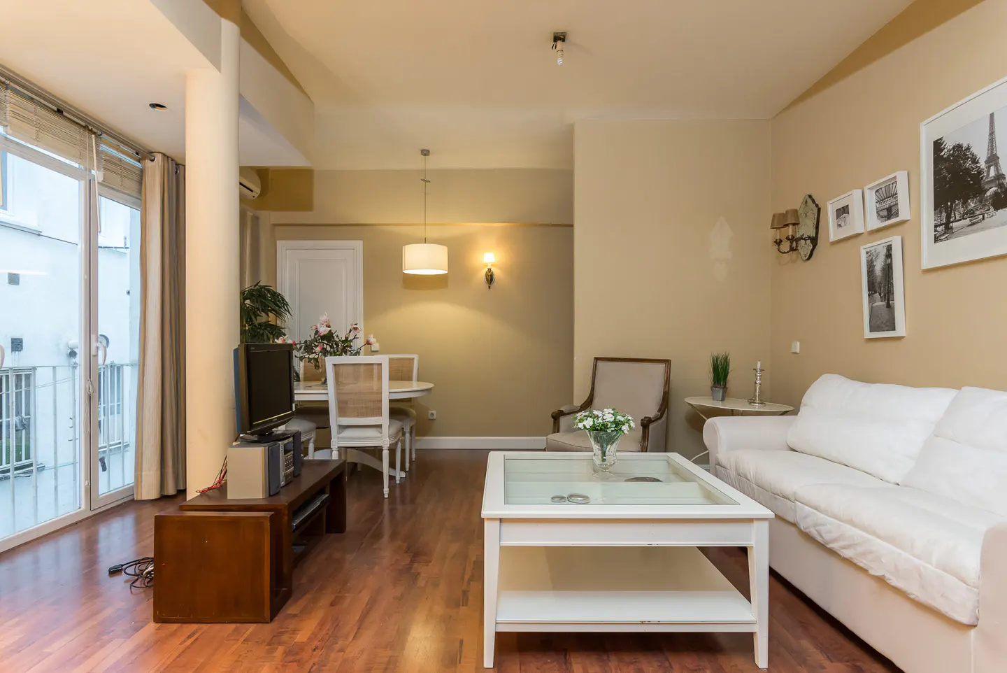 Living room with hardwood floors, white sofa, glass coffee table, dining table, and balcony with sliding glass doors.