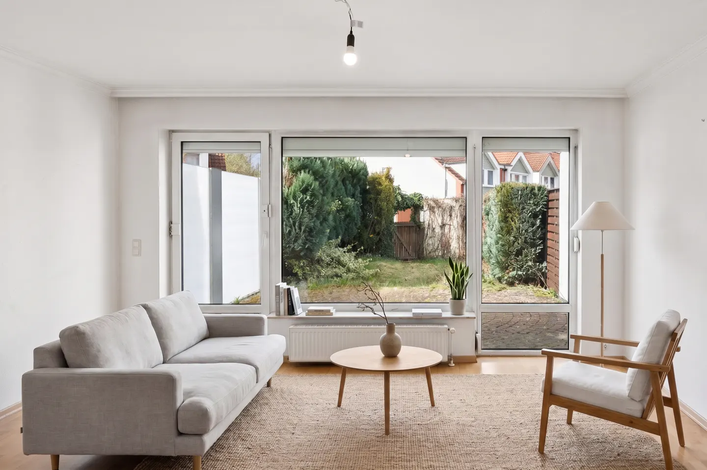 Bright living room with a gray sofa, wooden chair, and round table on a jute rug. Large windows overlook a green backyard.