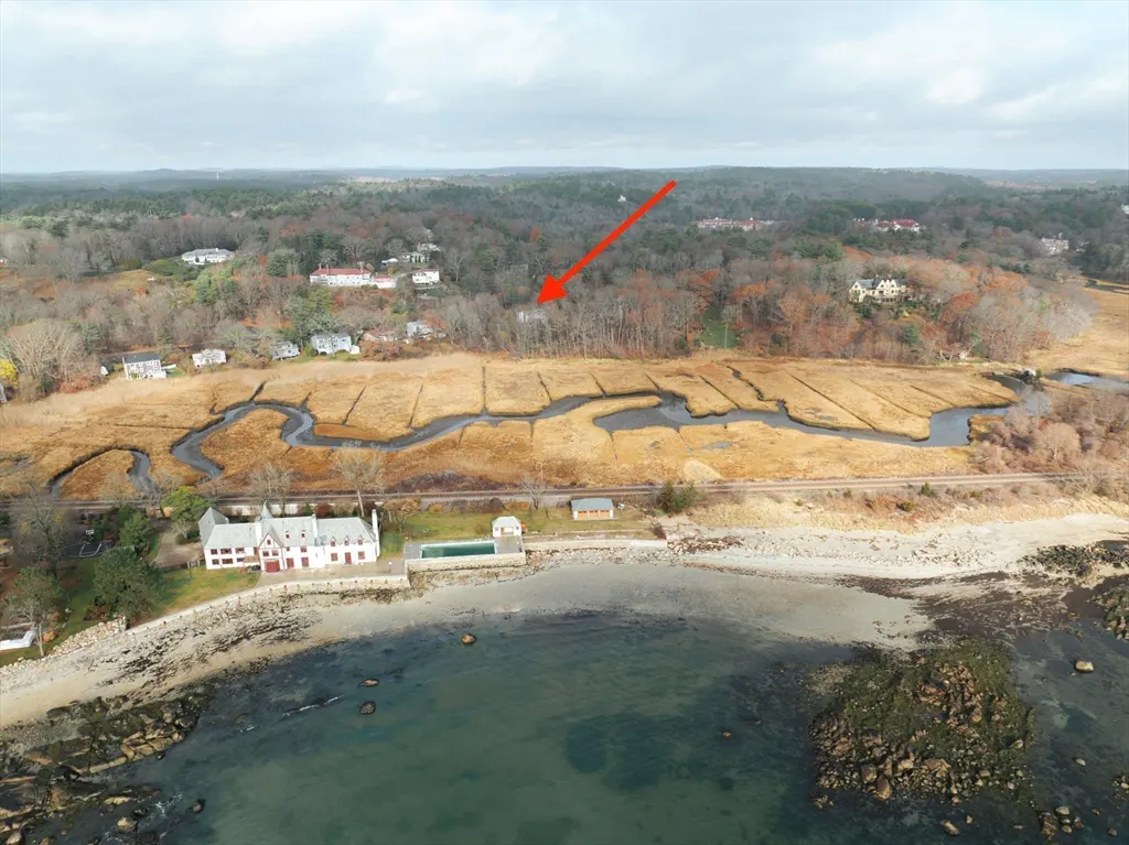 Aerial view of a coastal property with a large white house, pool, and beach, with an arrow pointing to a house in the distance.