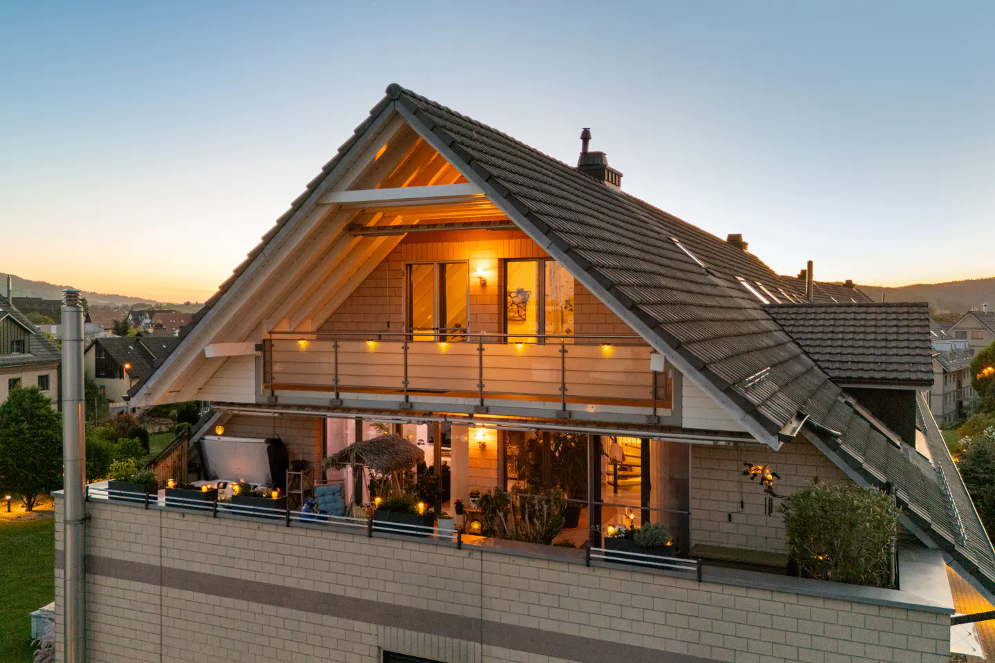 Exterior shot of a modern house with a large rooftop terrace at dusk. Warm lights glow from the windows and terrace.