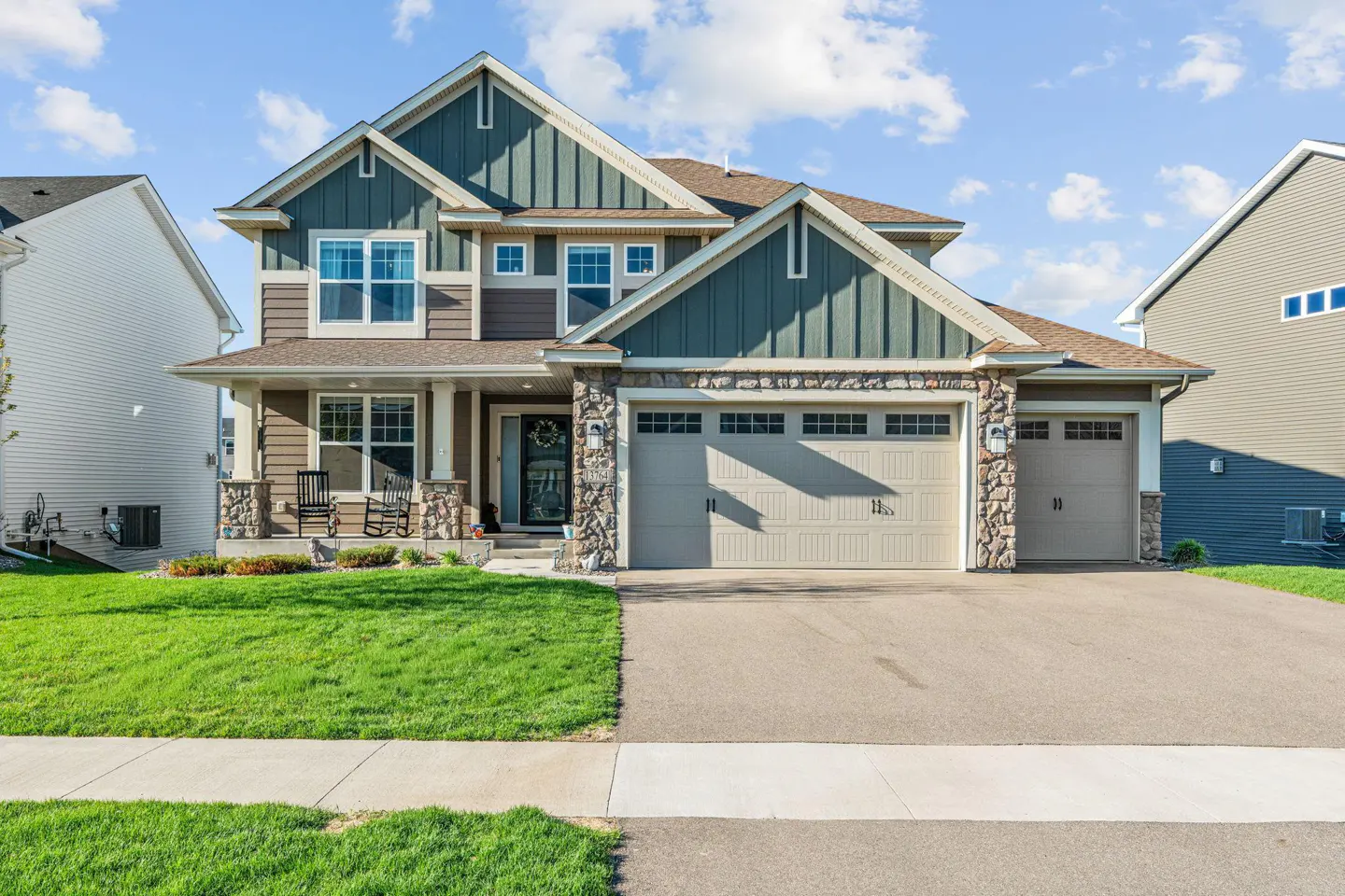 Two-story house with brown and green siding, stone accents, and a three-car garage. Lawn and driveway in front.