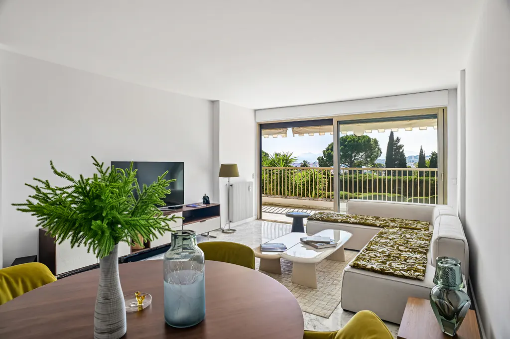 Bright living room with a brown table, green chairs, and a white sofa. Balcony doors open to a view of trees and mountains.