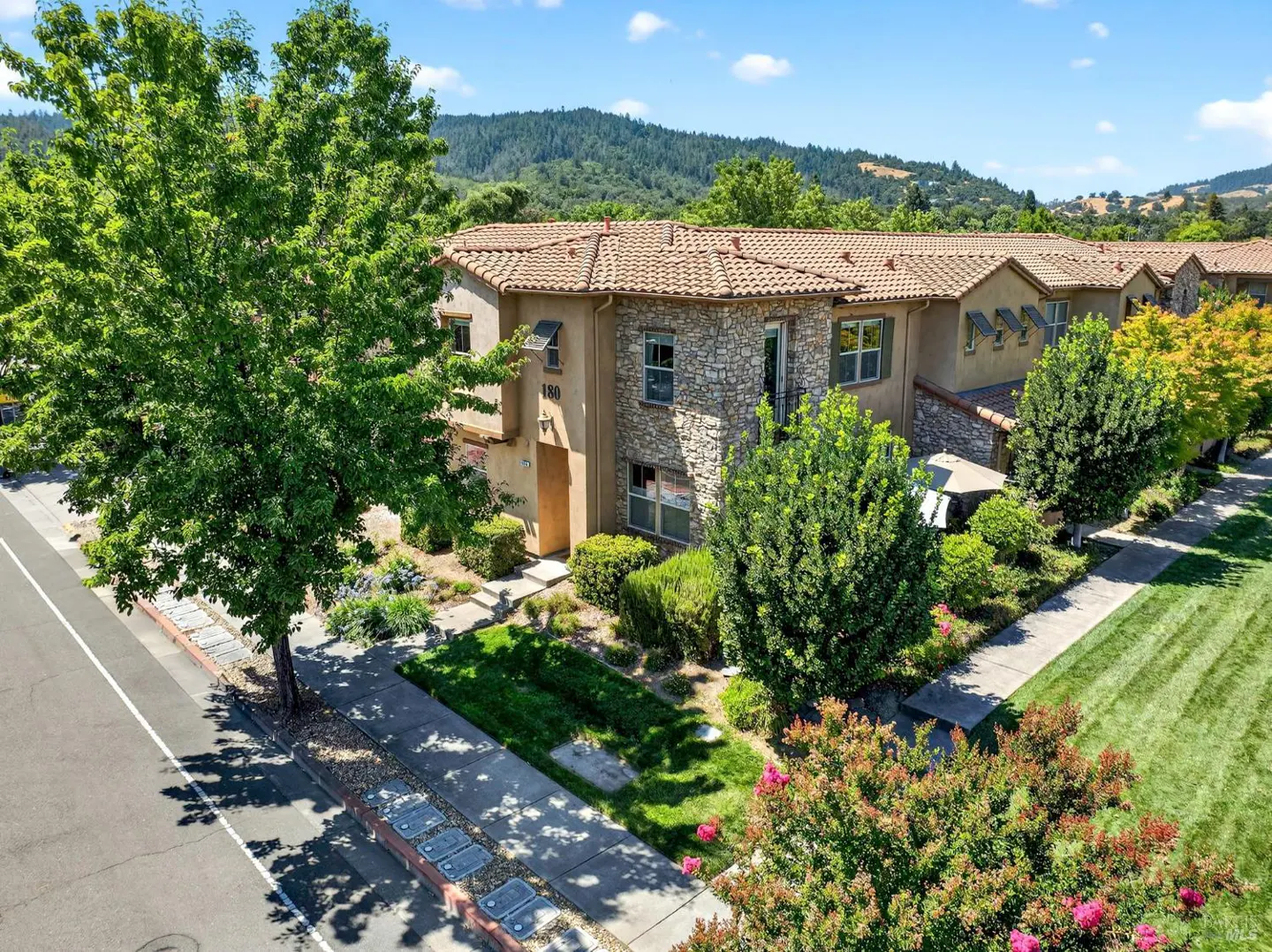 Two-story stone and stucco townhouse with a tile roof, surrounded by lush trees and landscaping under a blue sky.