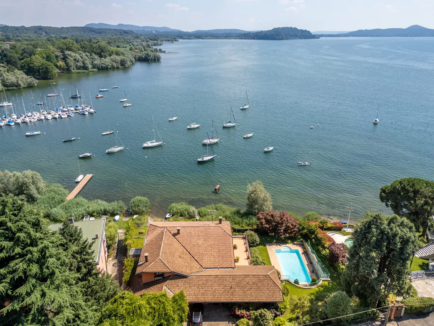 Aerial view of a lakeside property with a red-tiled roof, a blue swimming pool, and many sailboats on the water.