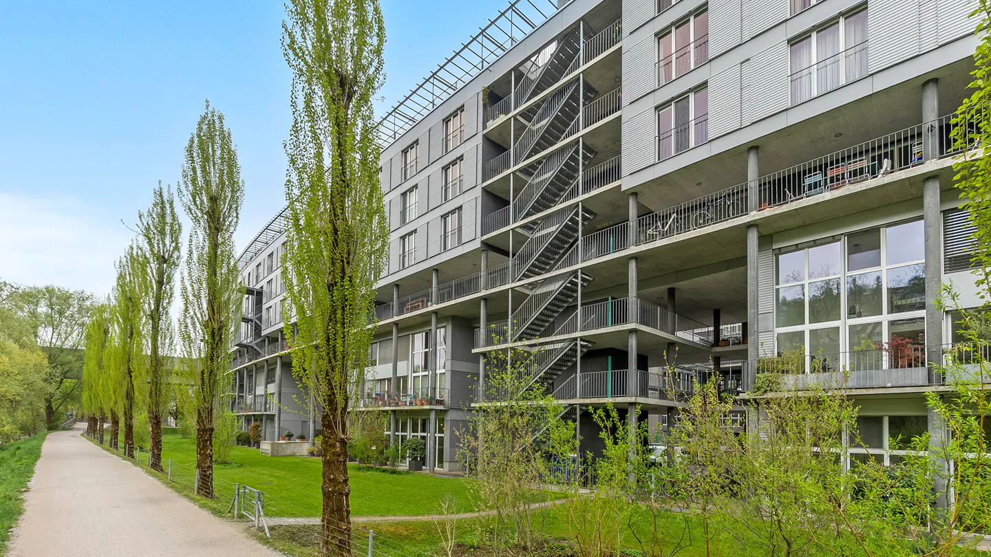 Exterior of a modern gray apartment building with balconies and metal stairs, next to a path lined with tall, thin trees.