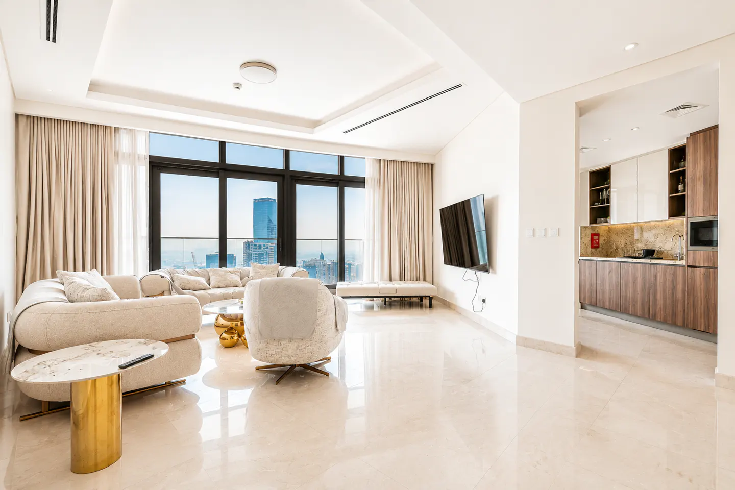 Bright, modern apartment living room with a beige sofa, chair, and city view. A TV hangs on the wall near the kitchen.