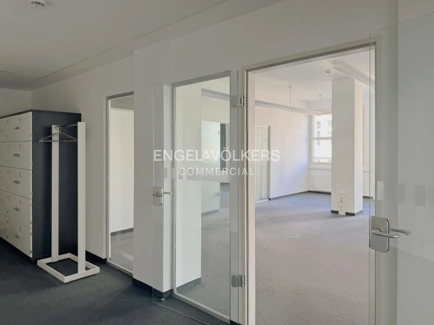 Bright office space with gray carpet, white walls, and glass-walled rooms. Lockers and a coat rack are on the left. Engel & Völkers Commercial logo on glass.