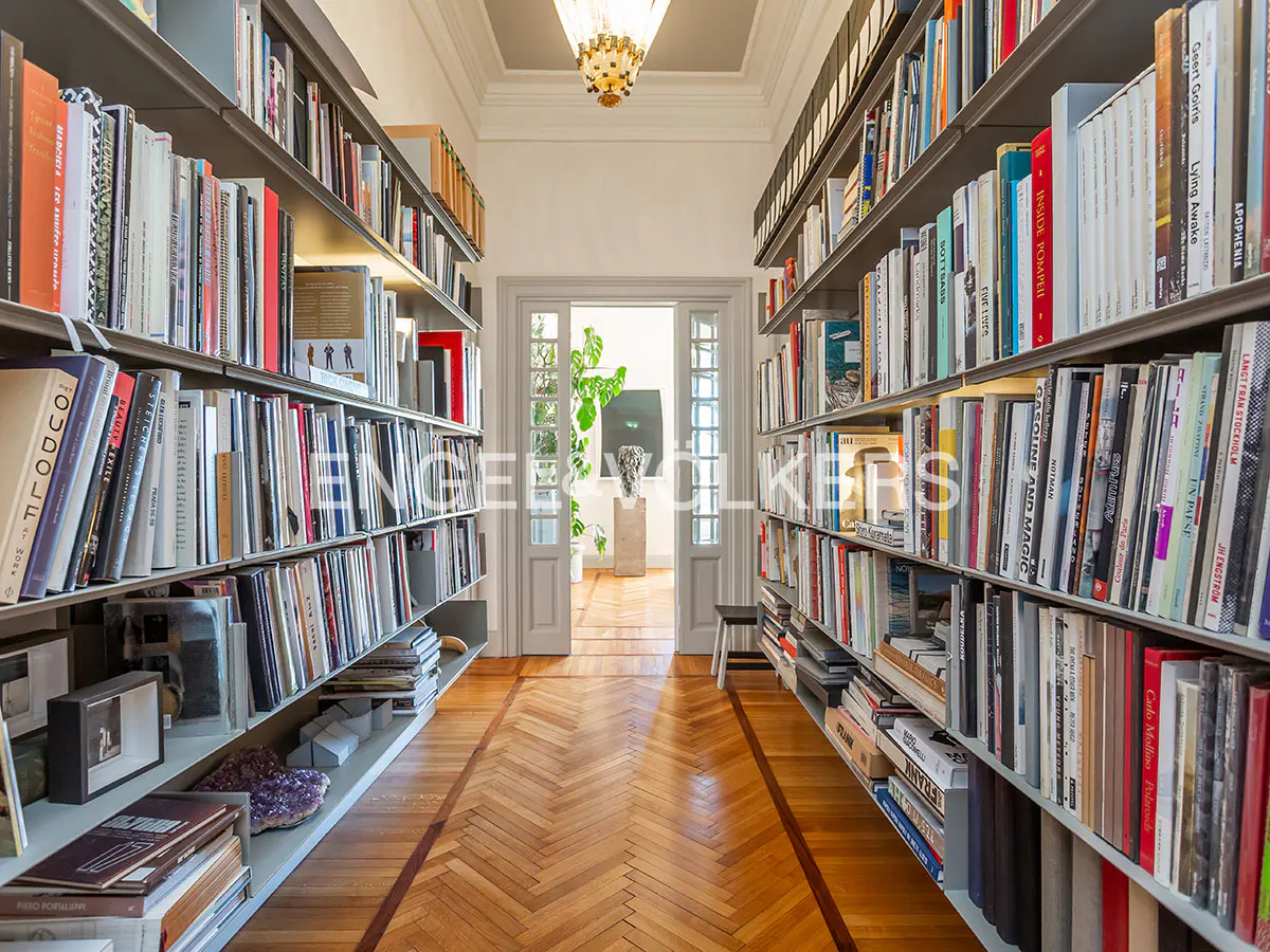 Hallway with floor-to-ceiling bookshelves filled with books, a herringbone wood floor, and a doorway to a bright room.