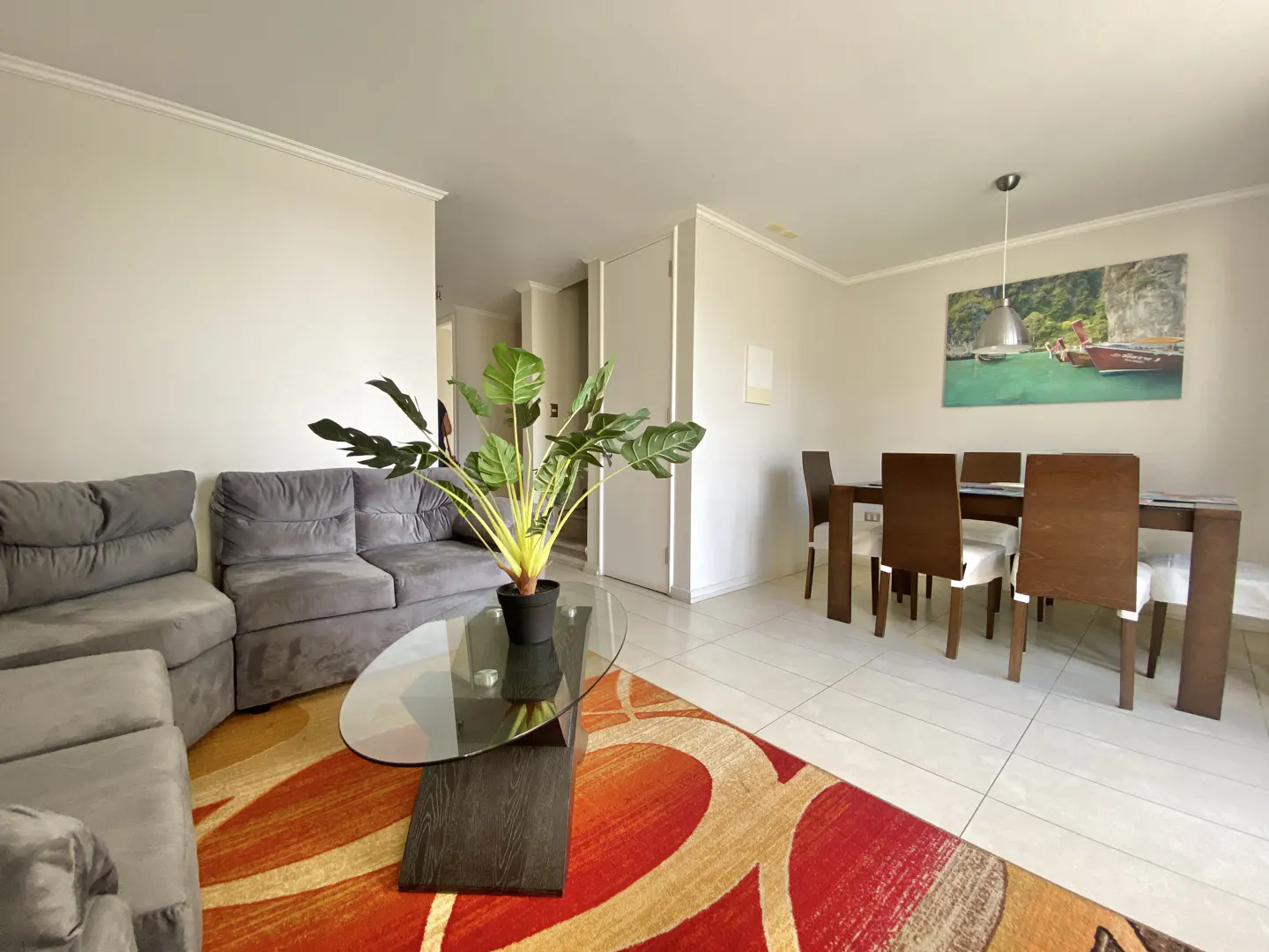 Living and dining area with gray sofa, glass table, and wooden dining set on a red and beige patterned rug. A tropical painting hangs above the table.