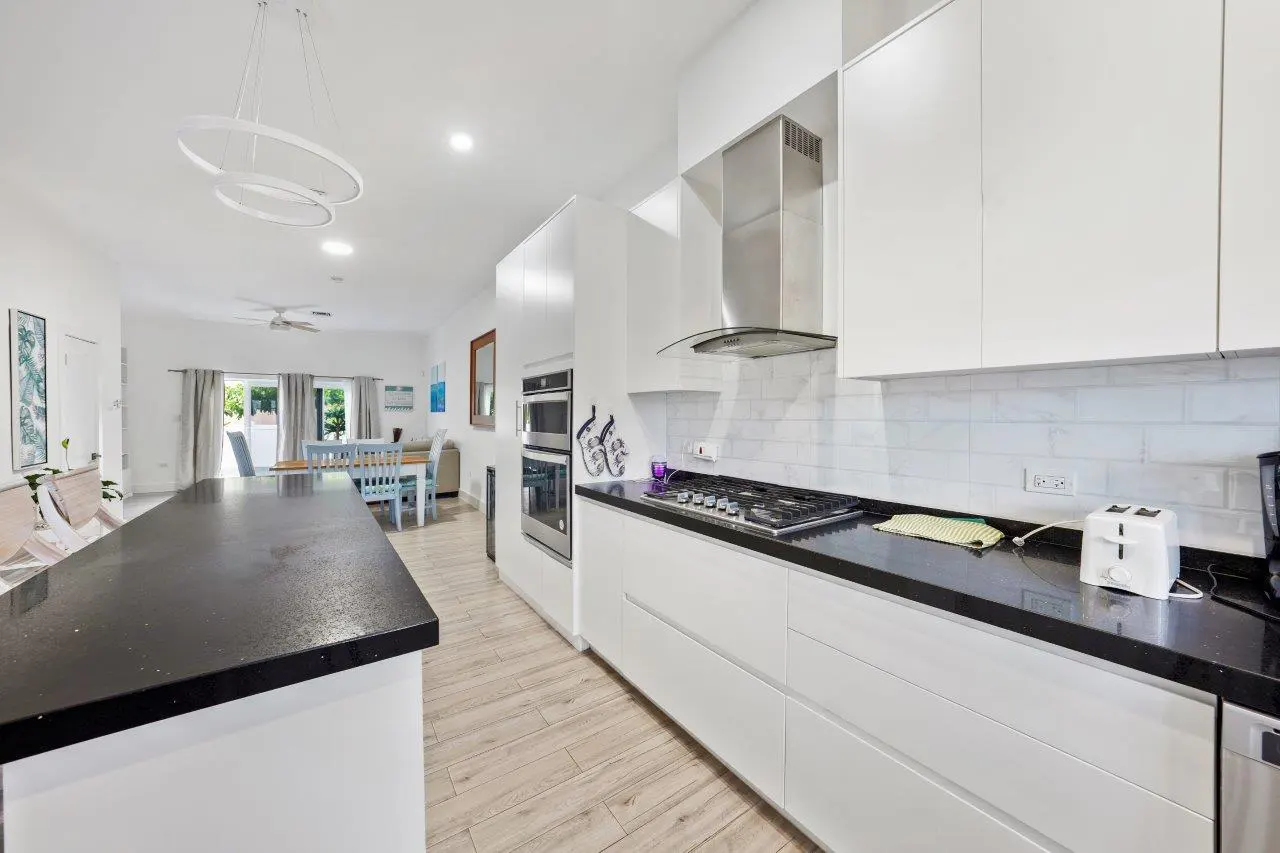 A bright, modern kitchen with white cabinets, black countertops, and stainless steel appliances. A large island extends into an open dining area.