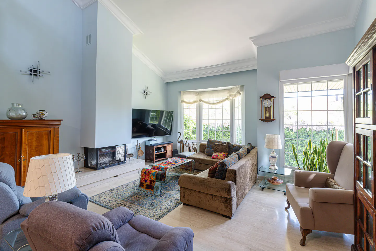 Living room with blue walls, a brown sectional sofa, a fireplace, and large windows. Two armchairs are in the foreground.