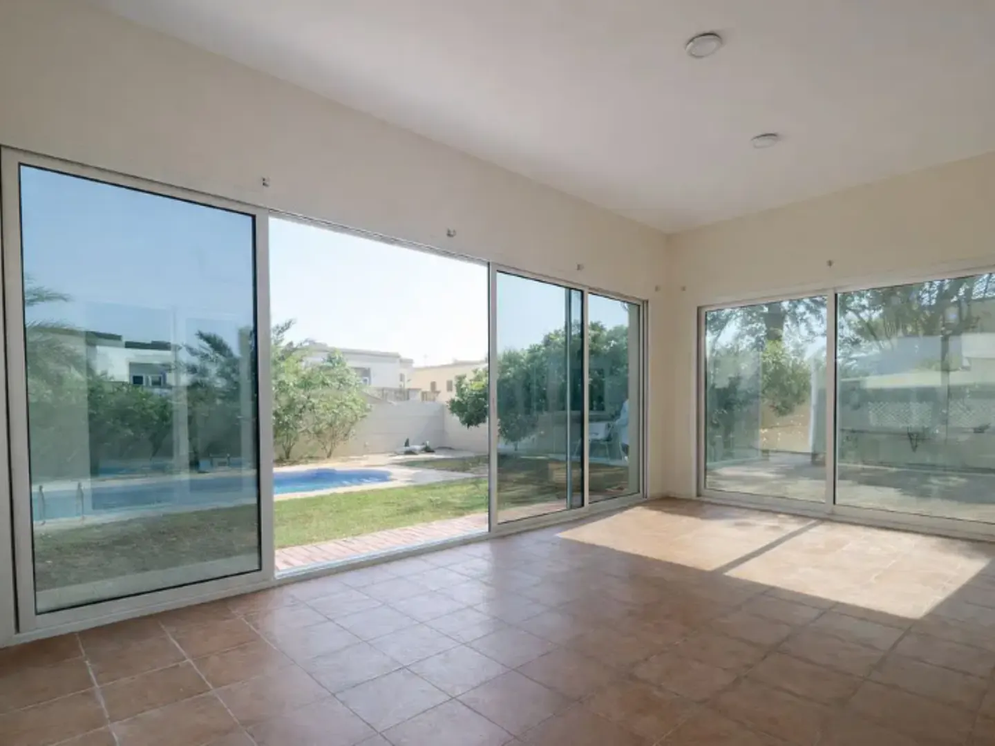 An empty room with tan tile floors and large sliding glass doors that open to a backyard with a pool.
