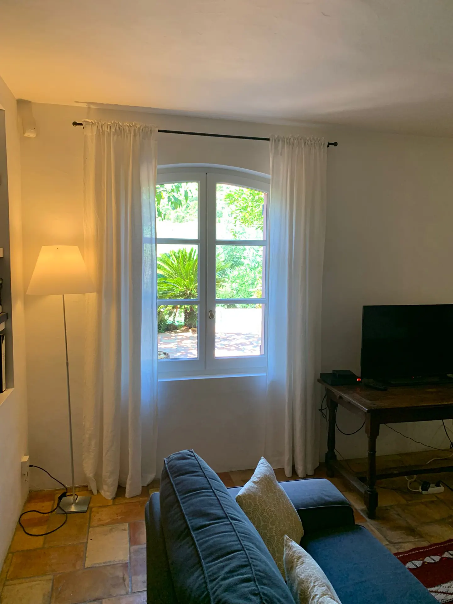 Living room with a blue sofa, a floor lamp, and a window with white curtains overlooking a garden.