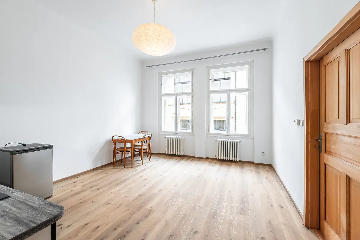 Bright, empty room with white walls, wood floors, and two windows. A small table and chairs sit near the windows, and a mini-fridge is in the corner. A wooden door is on the right.