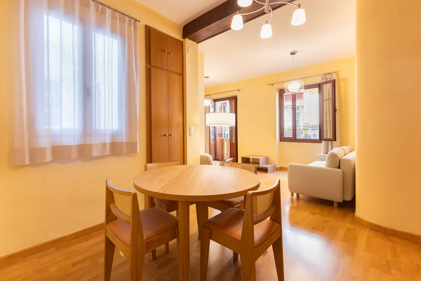 Bright living area with a round wooden dining table, chairs, and a cozy sofa in a warm, yellow-toned open-plan apartment.