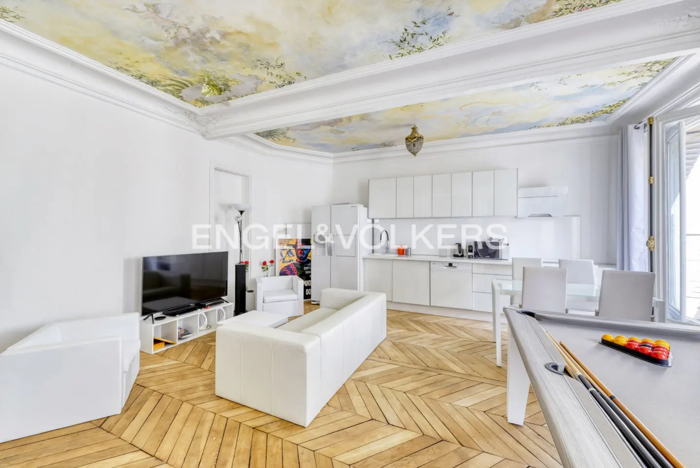 Bright, open apartment with white furniture, herringbone wood floors, and a painted ceiling mural. A pool table sits near a modern, white kitchen.