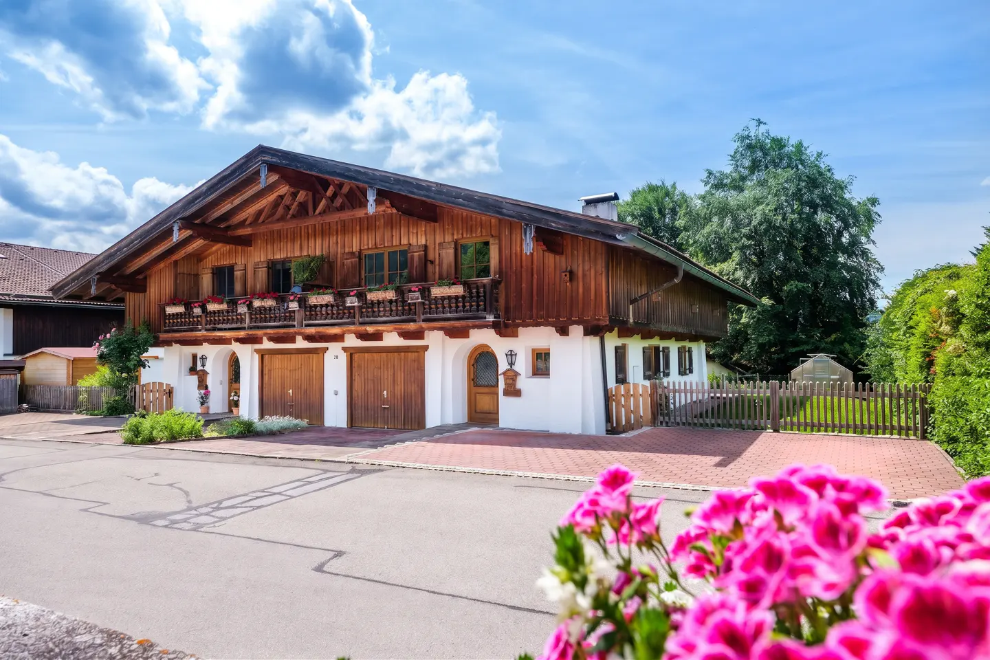 Traditional Bavarian-style house with wood siding, a balcony, and two garage doors, framed by bright pink flowers.
