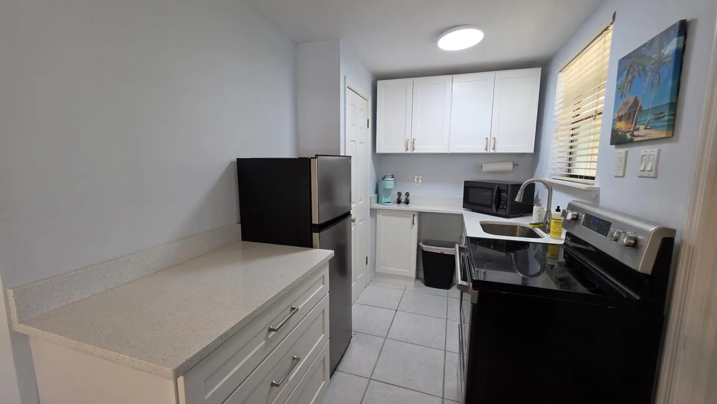 A compact kitchen with white cabinets, black appliances, and light blue walls. A tropical painting hangs near a window with blinds.