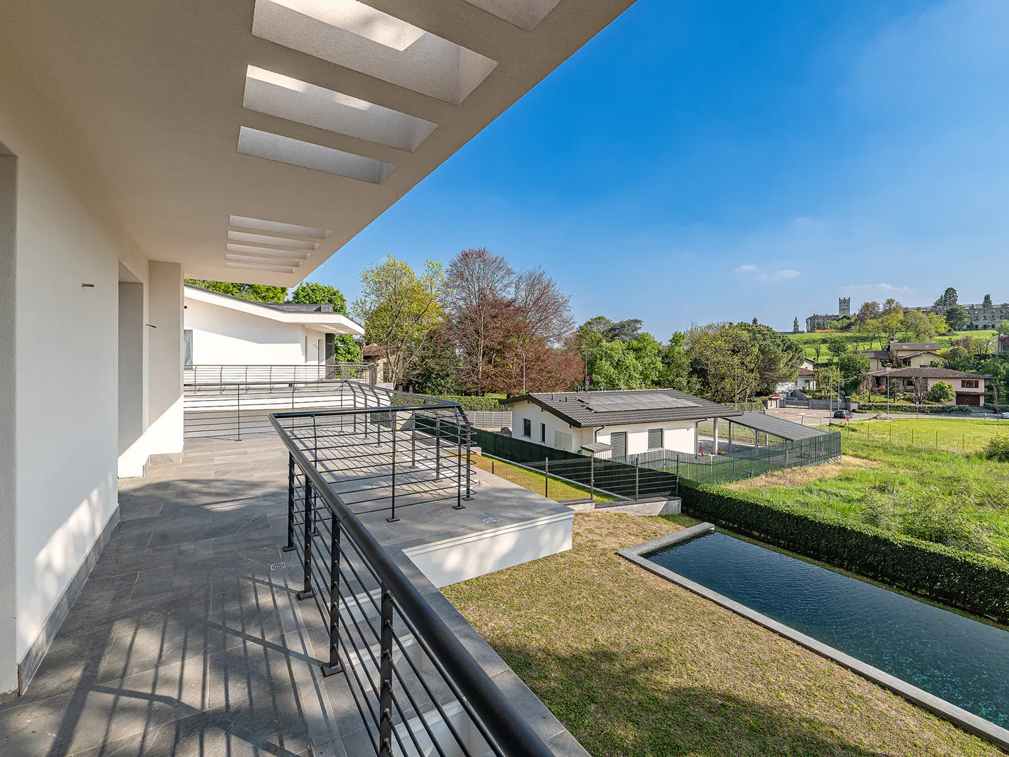 View from a modern home's balcony with black railings, overlooking a pool, green lawn, and houses under a blue sky.