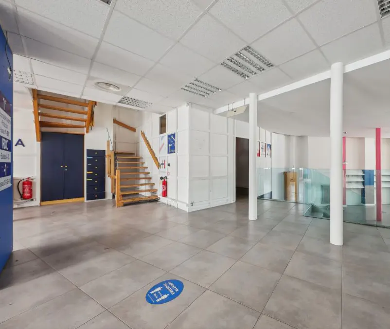 Empty commercial space with gray tile floor, white walls, and stairs. Blue signage and fire extinguishers are visible.