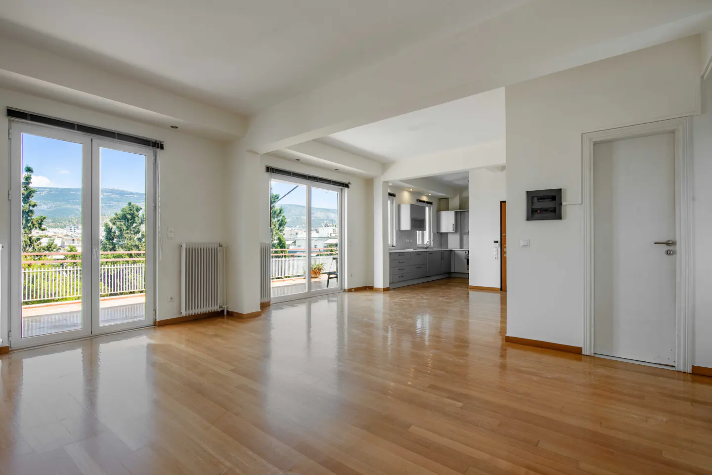 Bright, open-concept apartment with hardwood floors, white walls, and large windows leading to balconies with city views. A modern gray kitchen is visible.
