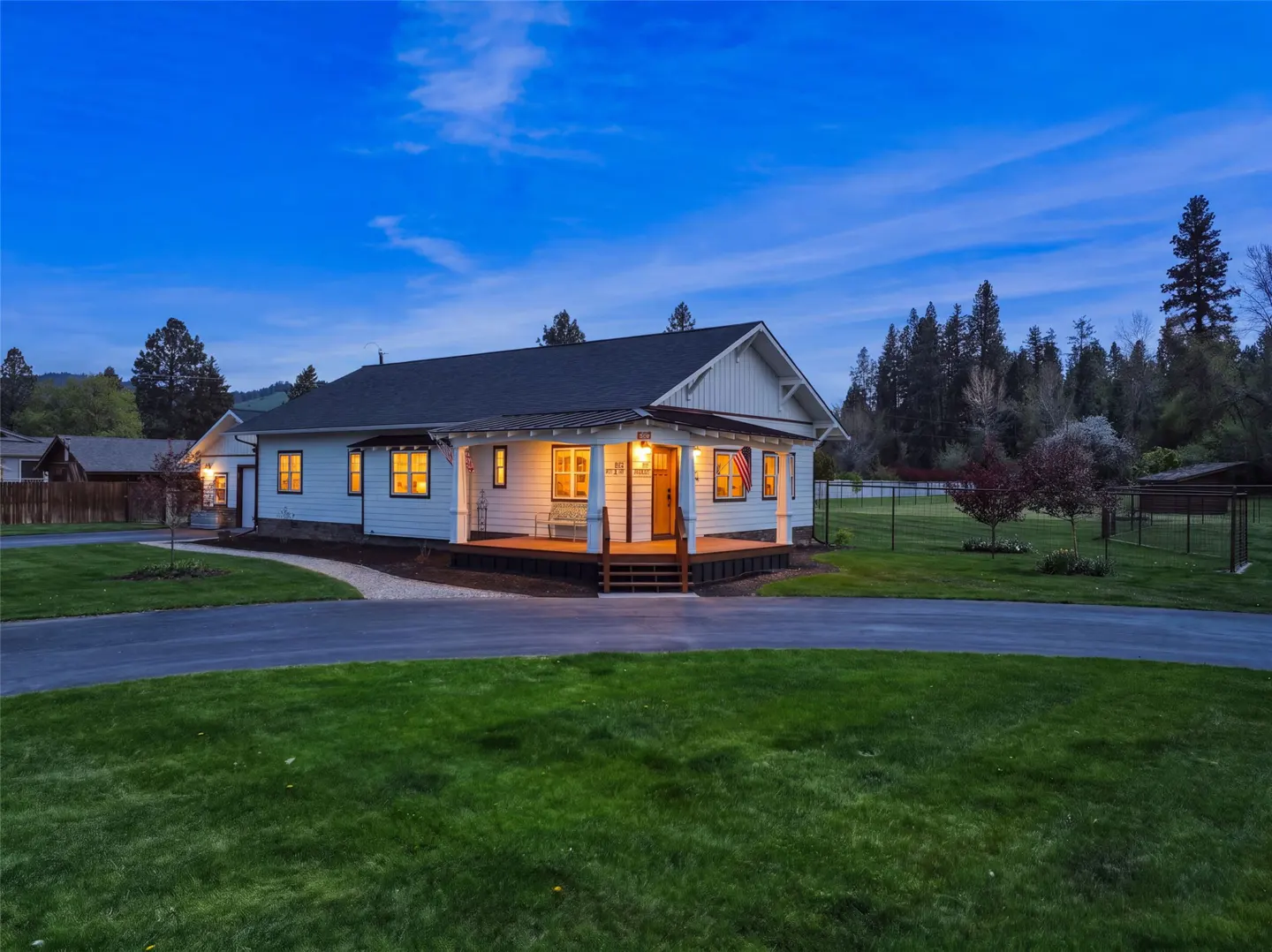 Exterior view of a white farmhouse with a dark roof, a porch, and a green lawn at dusk.